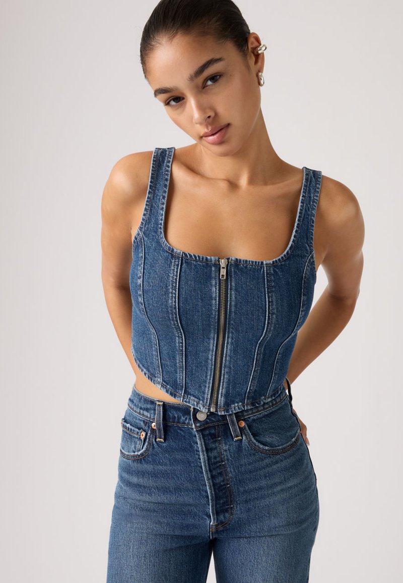 Νεαρή γυναίκα με τα μαλλιά της χτενισμένα προς τα πίσω φοράει μια εφαρμοστή τζιν crop top με φερμουάρ και ψηλόμεσα μπλε τζιν παντελόνια μπροστά σε απλό φωτεινό φόντο.