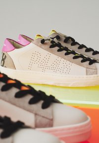 Paire de baskets beige et grises avec des lacets noirs et des languettes roses au talon, posées sur une surface réfléchissante colorée.