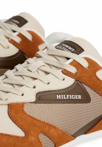 Baskets beige et marron présentant un mélange de textile, daim et cuir avec des panneaux en maille, un logo bien visible et des éléments de design contrastants.