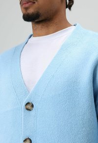Cardigan bleu clair en tissu texturé, doté d'un col en V et de trois boutons marron sur le devant. Porté par-dessus une chemise blanche.