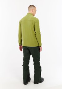 Man die met zijn rug naar de camera staat, gekleed in een lichtgroene langemouwen top, donkere groene broek en zwarte laarzen, tegen een eenvoudige witte achtergrond.