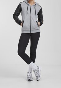 Sudadera gris con cremallera y mangas acolchadas negras, acompañada de leggings negros y zapatillas deportivas grises. Fondo liso.
