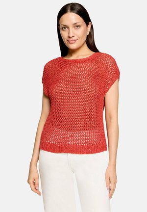 Femme aux cheveux raides et foncés portant un pull rouge en maille ajourée à manches courtes et un pantalon blanc, debout devant un fond clair uni.