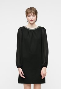 Young woman with short brown hair wearing a black long-sleeve dress with sheer sleeves and a beaded neckline, standing against a white background.