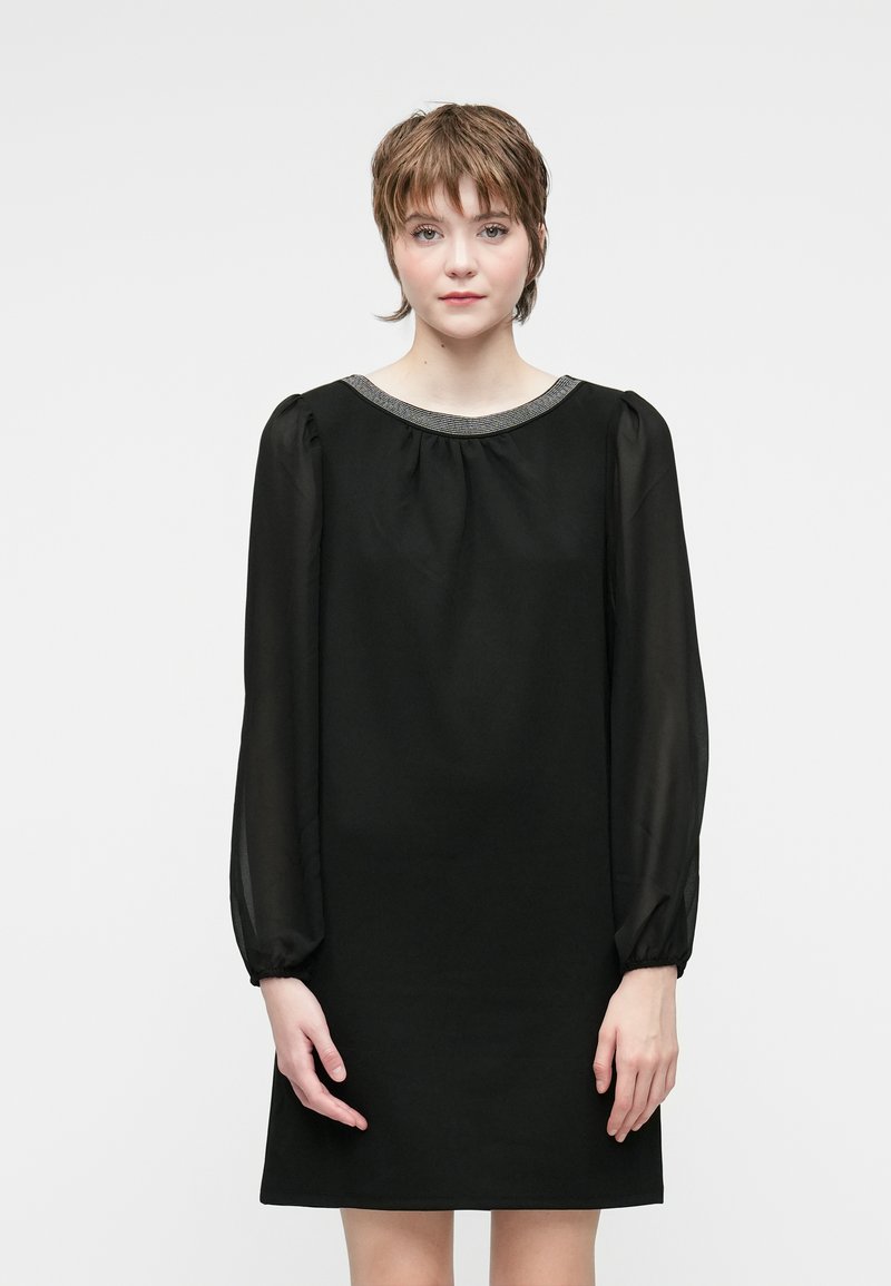 Young woman with short brown hair wearing a black long-sleeve dress with sheer sleeves and a beaded neckline, standing against a white background.