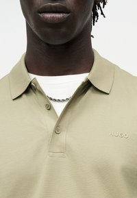 Κοντινό πλάνο ενός ατόμου που φορά ένα ανοιχτό ελαιώδες polo HUGO πάνω από ένα λευκό t-shirt με ασημένιο κολιέ αλυσίδα.