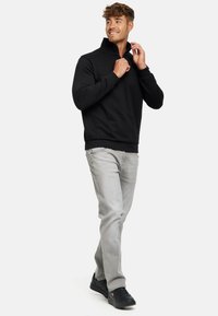 Pull noir à manches longues avec un col montant, associé à un jean gris clair et des baskets noires. Le modèle se tient dans une pose neutre.