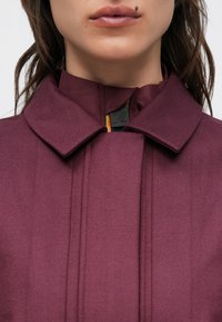 Maroon jacket with a high collar, featuring a zippered closure, vertical seams, and a textured fabric. Includes a colorful interior detail.