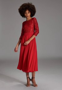 Afro frizurás nő piros flitteres felsőt és légies midi szoknyás ruhát visel, fekete magassarkú szandálban áll a szürke háttér előtt.