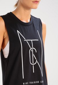 Mujer vistiendo una camiseta sin mangas negra de Nike Training Club con letras geométricas blancas y un sujetador deportivo blanco debajo.