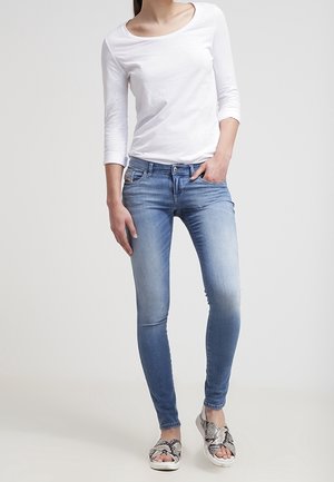 Femme portant un jean skinny bleu clair, une chemise blanche à manches longues et des sandales à enfiler à motif peau de serpent, debout avec une main dans la poche.