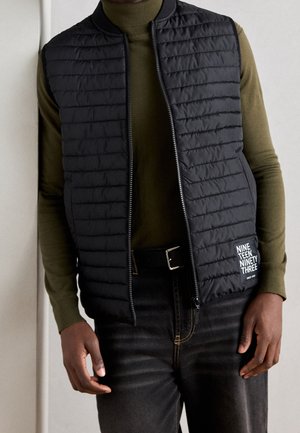 Uomo che indossa un gilet trapuntato nero sopra un dolcevita verde oliva, con jeans neri e cintura, in piedi davanti a uno sfondo chiaro.