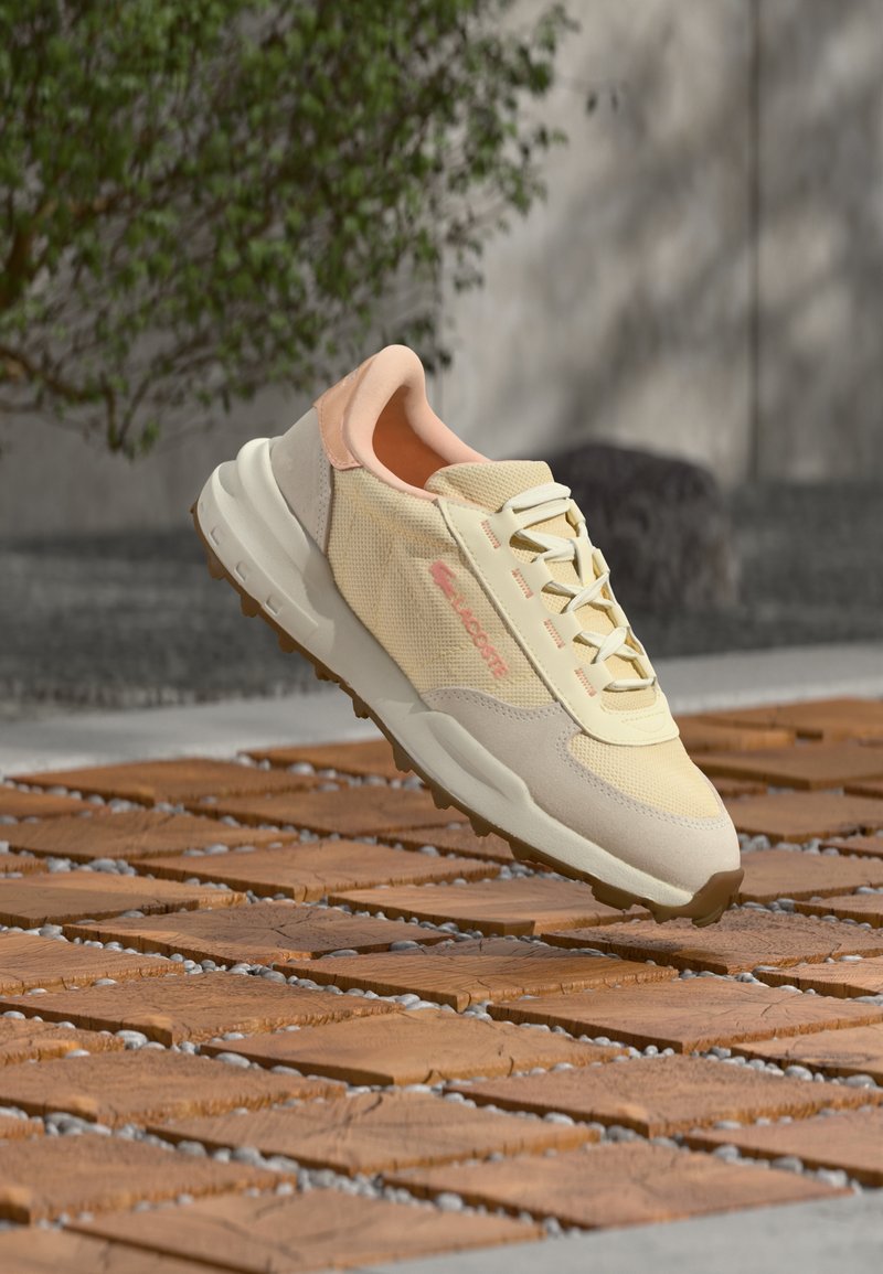 Sneaker in pale yellow and peach, featuring mesh and suede materials, a lace-up closure, and a thick, textured sole with rubber grip.