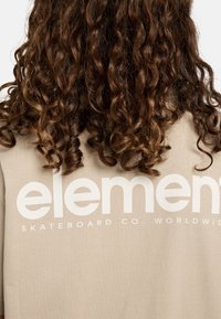 Bežové bavlněné tričko s velkým bílým nápisem na zádech "element Skateboard Co. WORLDWIDE". Viditelné kudrnaté hnědé vlasy.