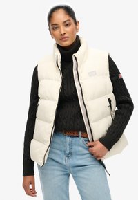 Smanicato imbottito beige con collo alto, chiusura con zip, tasche laterali, indossato sopra un maglione nero a trecce, abbinato a jeans azzurri.