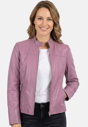 Lächelnde Frau mit welligem hellbraunem Haar trägt eine rosa, weiche Lederjacke mit Reißverschluss über einem weißen Hemd und schwarzen Hosen, die Hand in der Tasche.