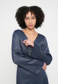 Rochie din satin albastru închis cu decolteu în V și mâneci lungi evazate; are un detaliu adunat în talie, textură netedă și o formă fluidă.