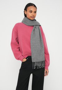 Szary wełniany szalik z postrzępionymi krawędziami, noszony na różowej bluzie. Modelka ma na sobie czarne spodnie i stoi na tle białego tła.