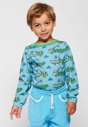 Ragazzo giovane con capelli biondi corti che indossa una maglietta blu con stampe di camion verdi e pantaloni azzurri con coulisse bianche.