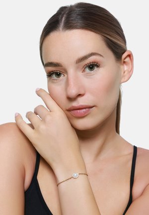 Bracciale in catena d'oro con un accento di perla iridescente rotonda, indossato su un polso nudo, con attenzione alla delicata texture della catena.