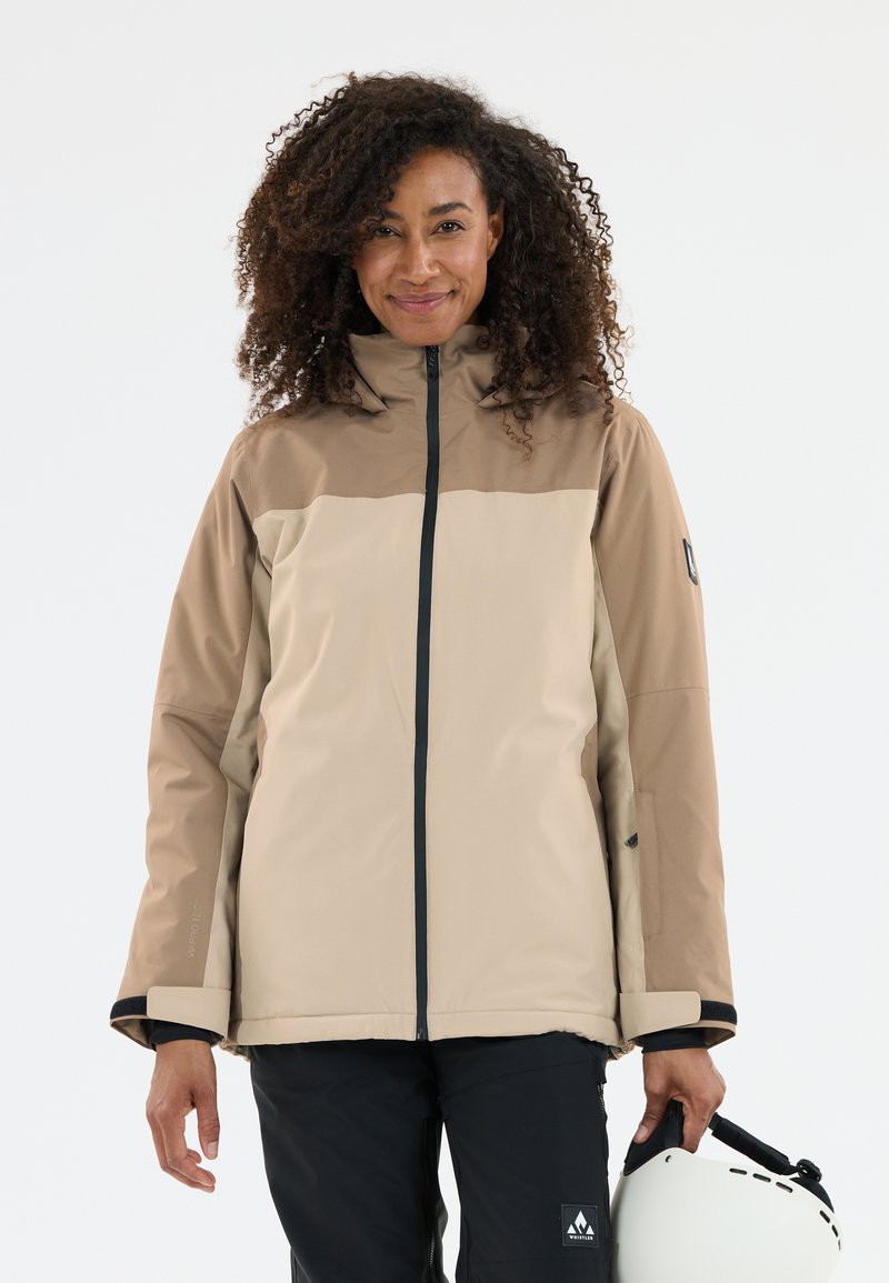 Tan og brun vandtæt jakke med høj krave, lynlås foran og sorte detaljer; kombineret med sorte bukser og en hvid hjelm.