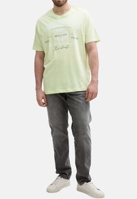 T-shirt en coton vert clair à manches courtes, avec texte et graphiques imprimés. Associé à un jean en denim gris et des baskets blanches.