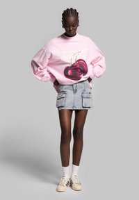 Sweatshirt overdimensionato rosa con grafica di ciliegie, abbinato a una gonna di jeans azzurra chiara con tasche cargo e sneakers beige con righe nere.