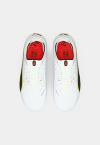 Scarpe da calcio bianche con dettagli neri e giallo brillante, caratterizzate da una tomaia testurizzata, lacci piatti e una soletta rossa con marchio.