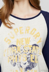 Witte en marineblauwe raglan shirt met een ronde hals. Bevat gele graphics met de tekst "SUPERDRY NEW YORK STATE" en een verouderd ontwerp.
