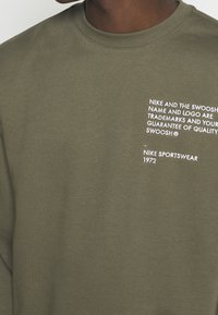 Hombre con sudadera verde oliva con el logo de texto blanco de Nike en el lado izquierdo del pecho que indica la marca registrada y la garantía de calidad.