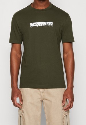 Mężczyzna ubrany w oliwkową koszulkę Calvin Klein New York i beżowe spodnie typu cargo, stojący na jednolitym tle.