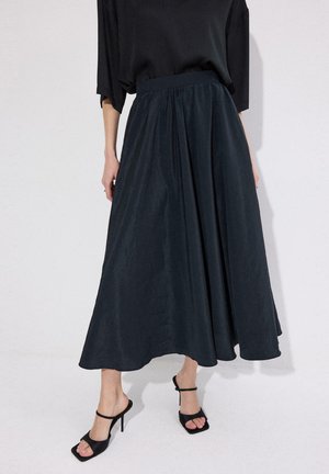 Femme portant une jupe noire midi trapèze et des sandales à talons hauts noires, debout devant un fond clair uni.