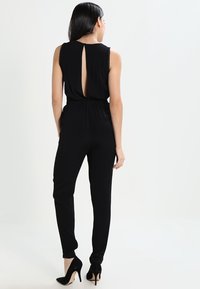 Černý overal bez rukávů, s otevřeným detailem na zádech, elastickým pasem a zúženými nohavicemi. Spárován s černými lodičkami na vysokém podpatku.