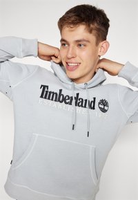 Felpa grigia chiara con tasca a marsupio, polsini a costine e grafica "Timberland Established 1973" in nero e crema. Materiale in cotone.
