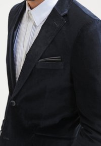 Blazer in velluto blu navy con un bottone e rever a scatto, abbinato a una camicia bianca plissettata, con particolare dettaglio nella tasca accentuato.