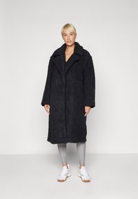 Cappotto teddy nero, in peluche, con colletto a rever, maniche lunghe e tasche laterali, indossato sopra leggings grigi e sneakers bianche.