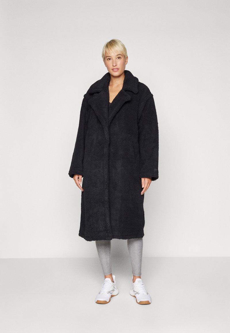 Cappotto teddy nero, in peluche, con colletto a rever, maniche lunghe e tasche laterali, indossato sopra leggings grigi e sneakers bianche.