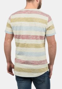 Tricou cu dungi în culoarea crem, cu dungi orizontale de diferite nuanțe de roșu, albastru și galben, având un gât rotund și mâneci scurte.