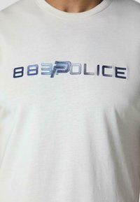 Majica s kratkim rukavima, svijetlosiva pamučna majica s plavim gradijentnim logotipom "883POLICE" preko prsa. Glatka tekstura, okrugli izrez.