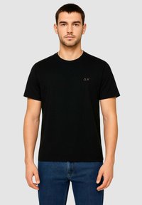 T-shirt nera in cotone con maniche corte, colletto rotondo e un piccolo logo dorato sul petto sinistro. Indossata con jeans denim blu.