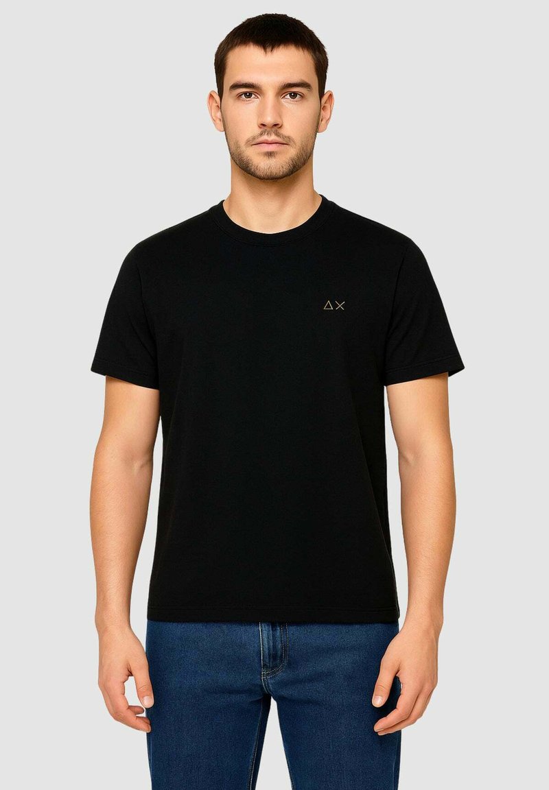 T-shirt nera in cotone con maniche corte, colletto rotondo e un piccolo logo dorato sul petto sinistro. Indossata con jeans denim blu.