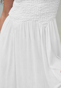 Robe blanche avec un corps smocké et une jupe fluide. Fabriquée dans un tissu léger, elle présente une texture douce et des détails froncés.