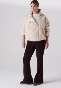 Fluffige cremefarbene Zip-Jacke mit hohem Kragen und weißer Fronttasche, kombiniert mit dunkelbraunen Schlaghosen und braunen Slippern.