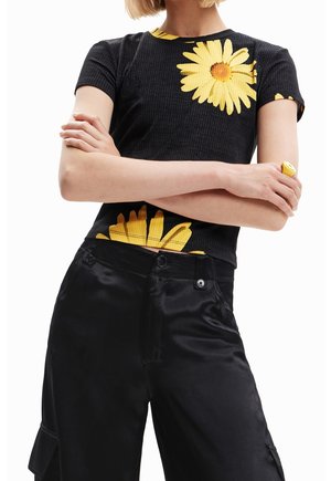 Mujer con los brazos cruzados que lleva una camiseta negra acanalada con grandes margaritas amarillas y pantalones anchos brillantes negros.