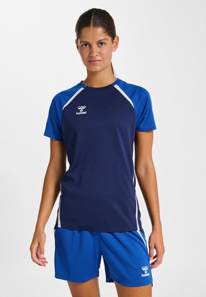 Kobieta w niebieskim i granatowym sportowym t-shircie oraz niebieskich szortach stoi na białym tle.