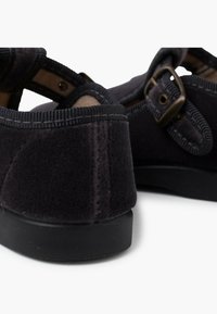 Scarpe in feltro nere con retro arrotondato, lati aperti e una tracolla regolabile con fibbia in bronzo. La suola è in gomma nera.