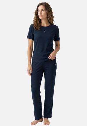 Kvinde med bølget hår står barfodet, iført en matchende marineblå kortærmet skjorte og bukser, kigger til siden mod en ensfarvet baggrund.