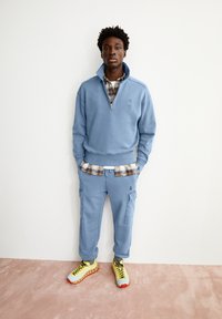 Pullover in pile azzurro chiaro con mezza zip e logo, abbinato a pantaloni cargo azzurro chiaro. Presenta una camicia a quadri sotto e scarpe da ginnastica vivaci.