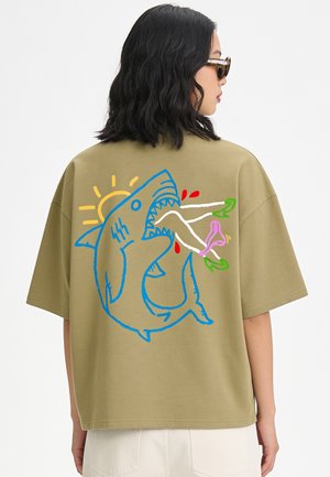 Frau trägt ein olivgrünes T-Shirt mit einer bunten Hai-Grafik, die ein Paar Beine mit roten Tropfen und eine gelbe Sonne im Hintergrund verschlingt.
