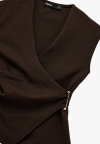 Top marrón sin mangas con un escote en V profundo, con un diseño envolvente y un detalle decorativo de pin dorado en un lado. Textura suave.